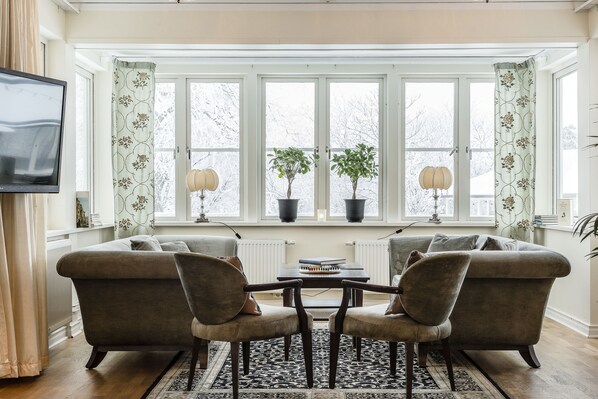 A living room with a sofa, two armchairs, a coffee table, and a TV on the wall. There are windows with curtains, a radiator, and two potted plants on the windowsill. - Såstaholm Hotel & Konferens (Taby)