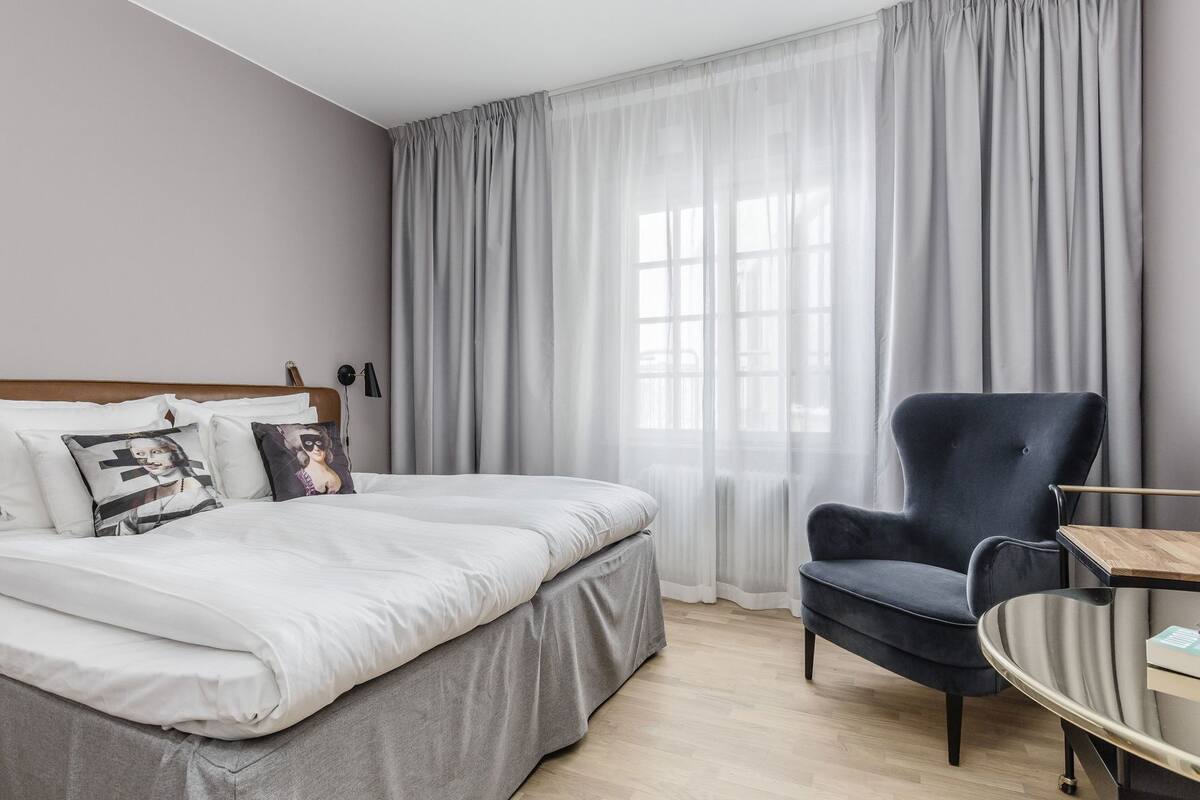 Un dormitorio con cama, una silla, una mesa y una ventana con cortinas.