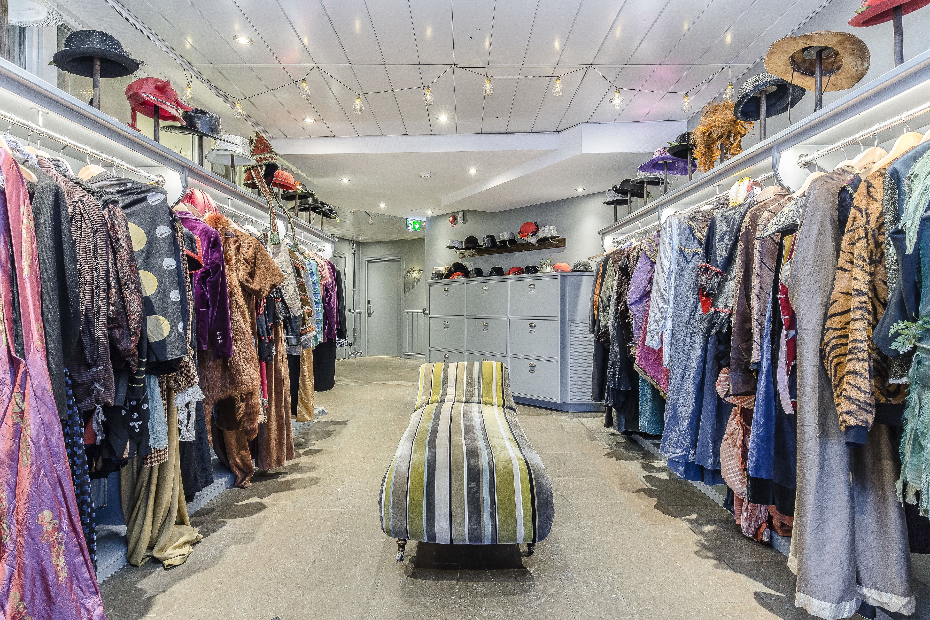Interior de un hotel boutique con una cama central, percheros con ropa a ambos lados y una variedad de sombreros exhibidos en estantes.