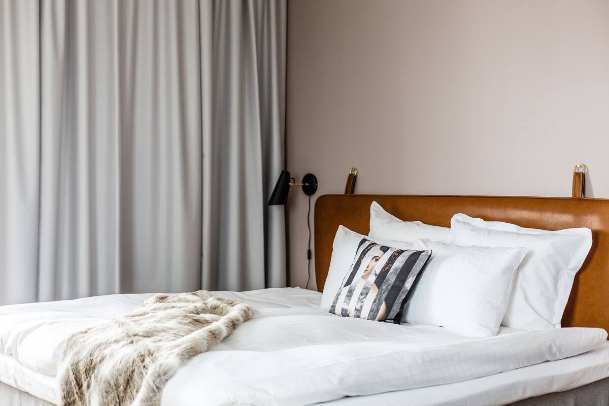 Una cama bien hecha con sábanas blancas y un plaid de piel, un cabecero de madera y una lámpara de pared.