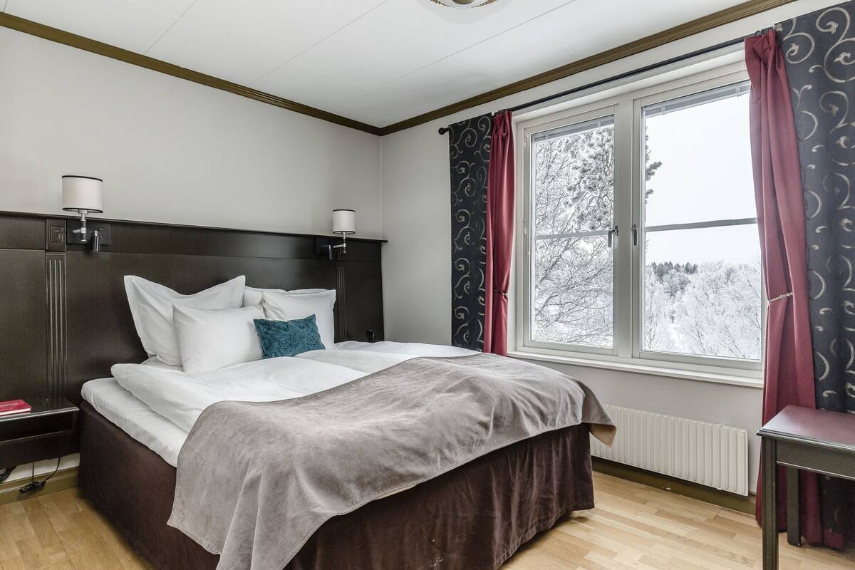 Habitación de hotel con cama, lámparas de noche, ventana con cortinas y vista a árboles.