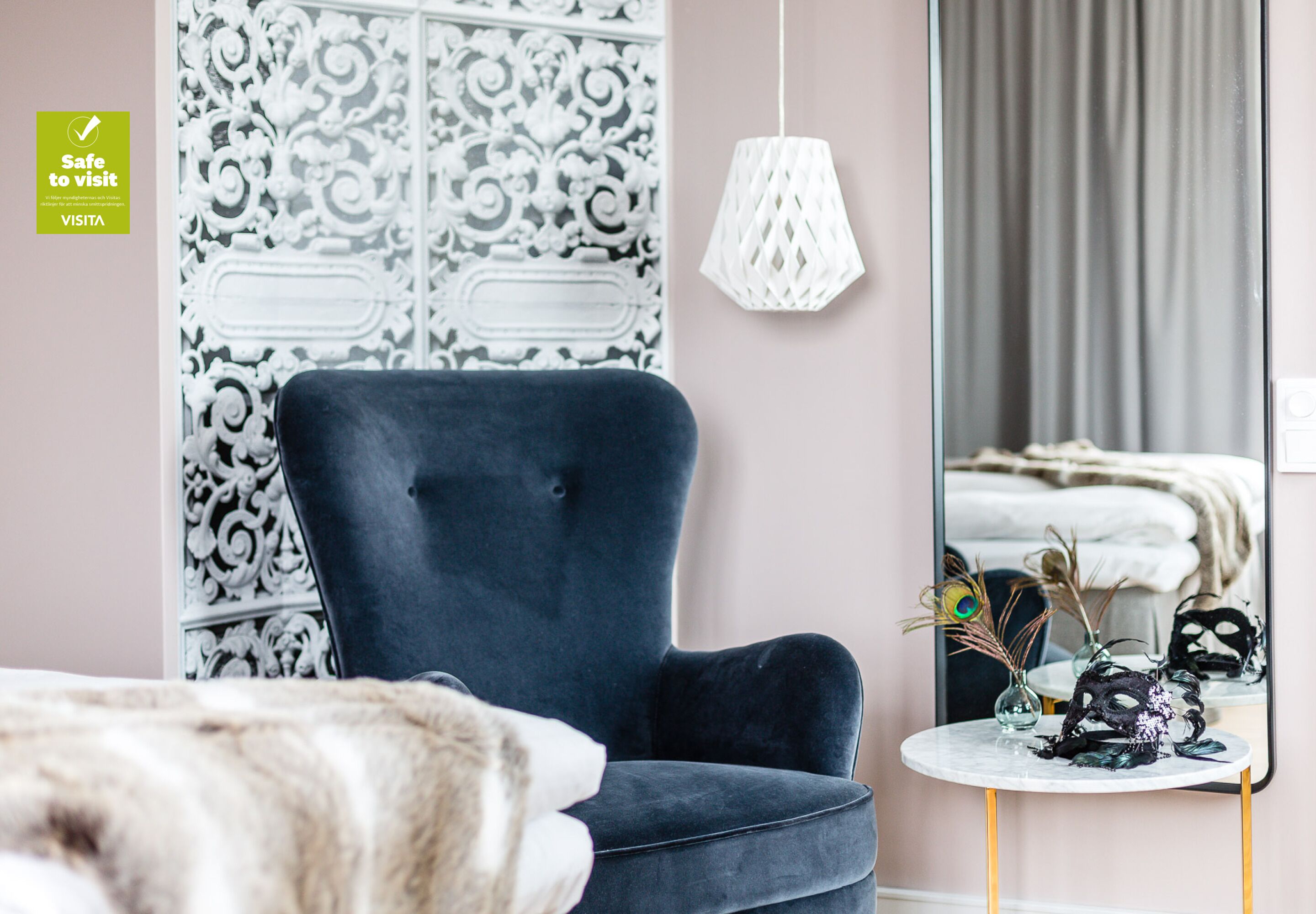 Una habitación moderna con un sillón azul oscuro, un espejo decorativo y una mesita redonda con un jarrón y una máscara.