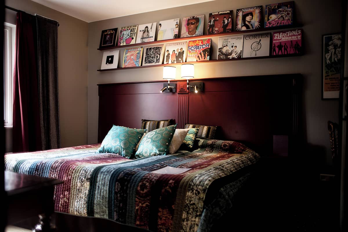 Una cama bien tendida con ropa de cama estampada y varios almohadones, una lámpara de pared y una serie de carteles enmarcados sobre la cama.