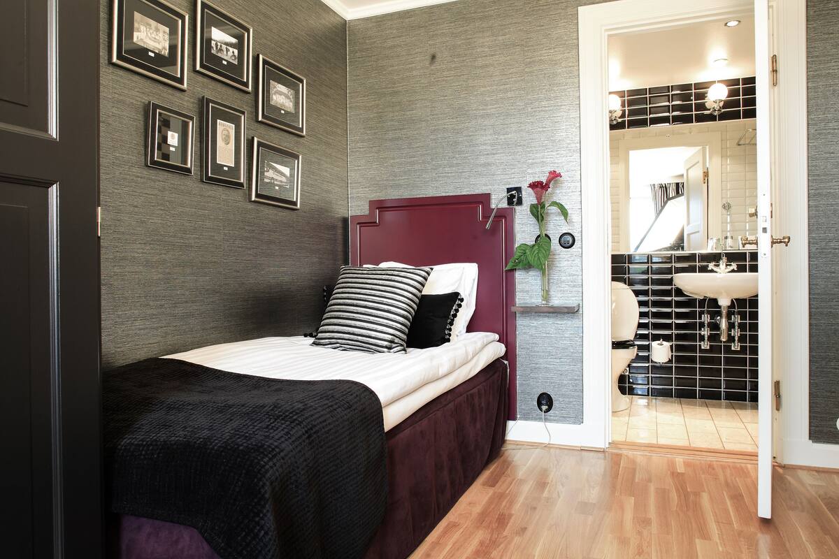 Un dormitorio con cama, cabecera, mesita de noche, estante de pared con cuadros enmarcados y un baño visible a través de una puerta abierta.