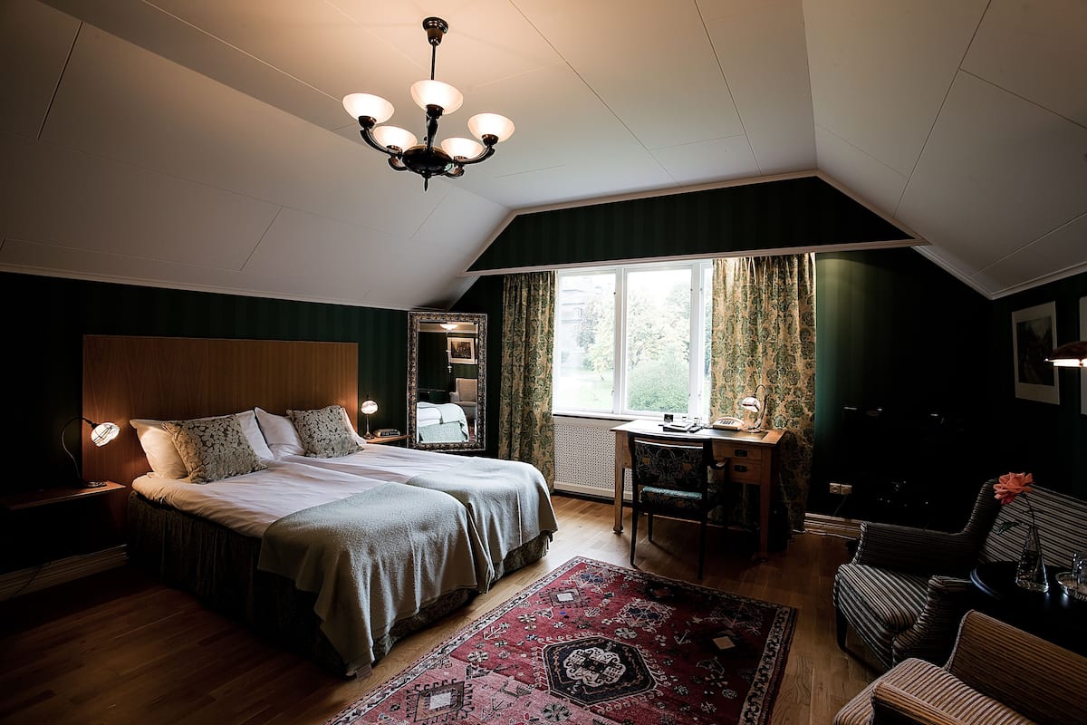Un dormitorio con una cama grande, un escritorio, una silla, una araña y un ventanal con cortinas.