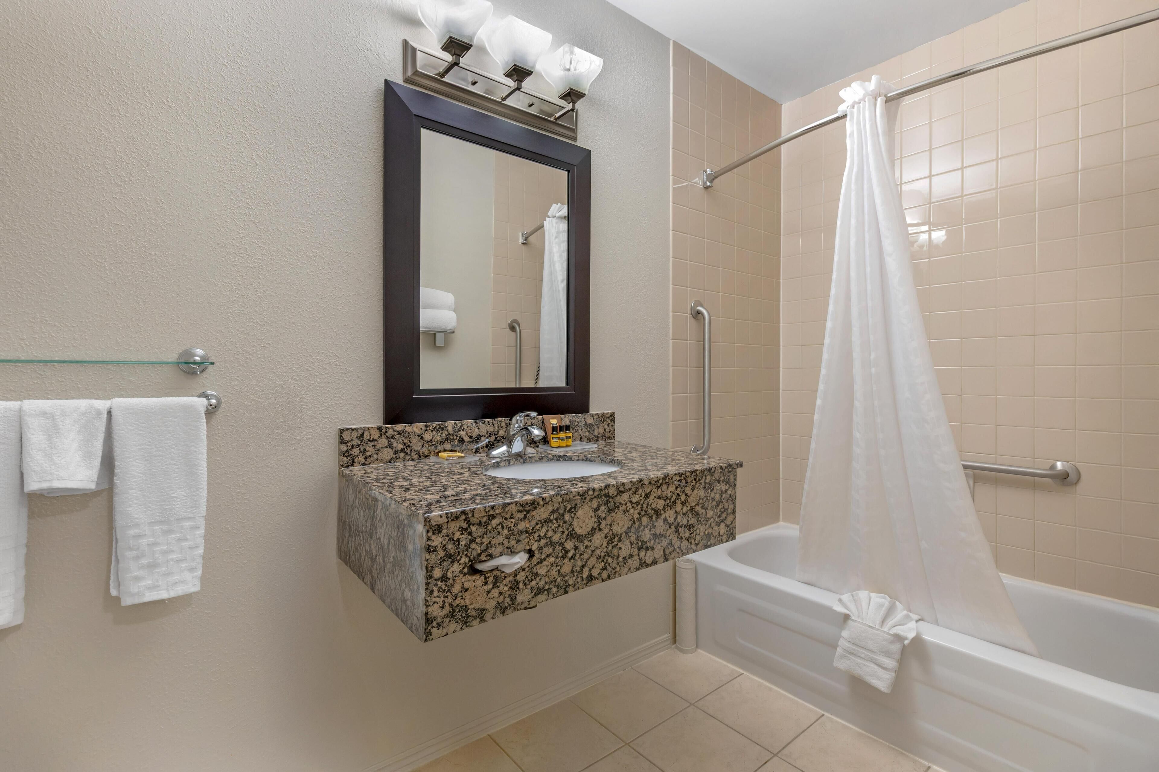 Une salle de bain avec un lavabo sur un plan de travail en granit, un miroir, un porte-serviettes, une douche avec un rideau et une baignoire.