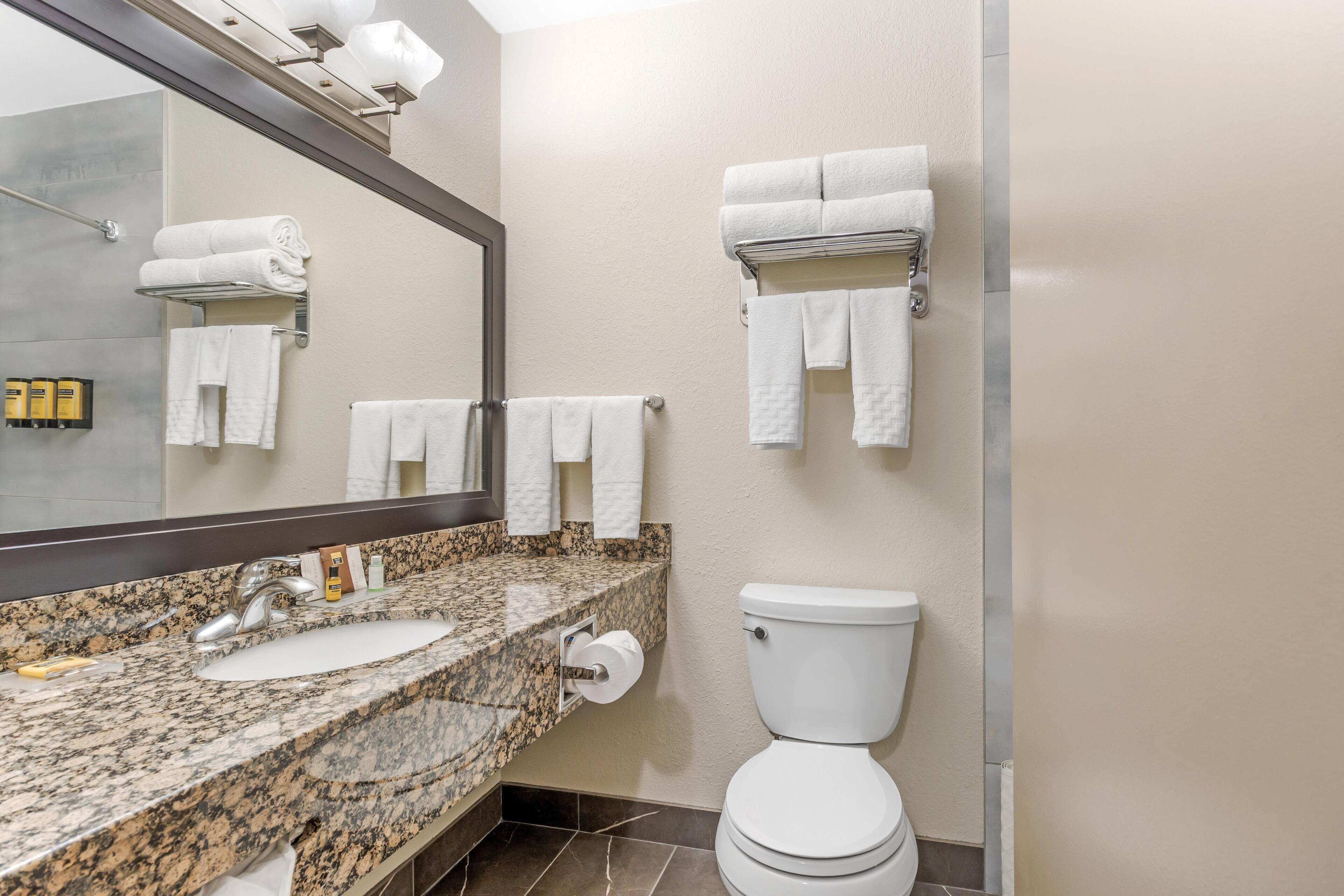 Un baño con encimera de granito, lavabo blanco y un inodoro. Hay varias toallas blancas colgadas en la pared y un espejo sobre el lavabo.