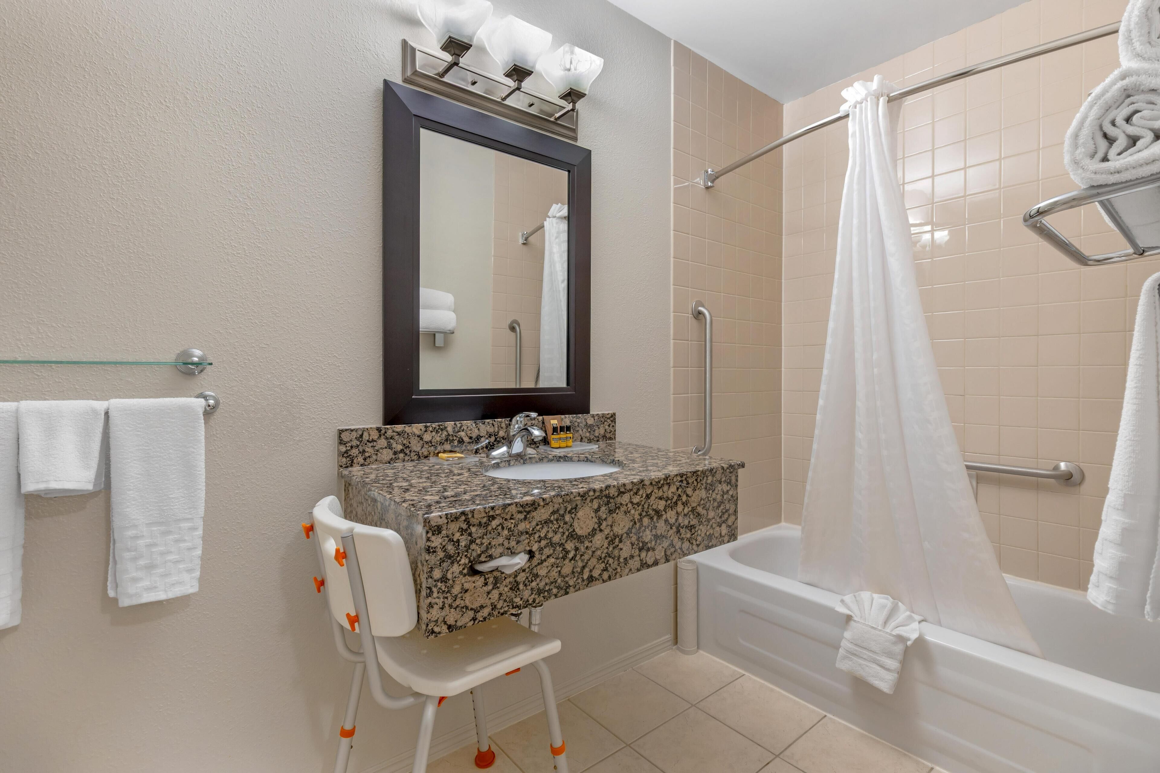 Un baño con lavamanos de granito, un espejo, una silla blanca, una bañera y una cortina de ducha.