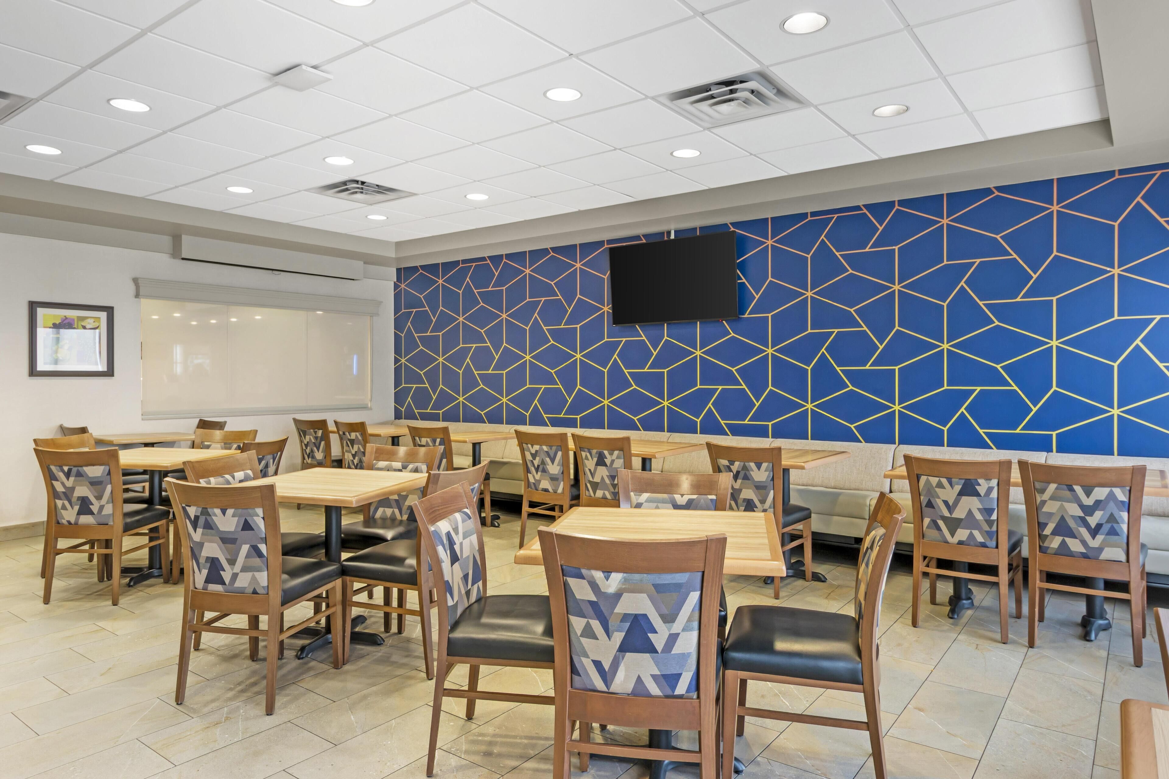 A dining area with wooden tables and chairs, a patterned wall, and a flat-screen TV mounted on the wall.