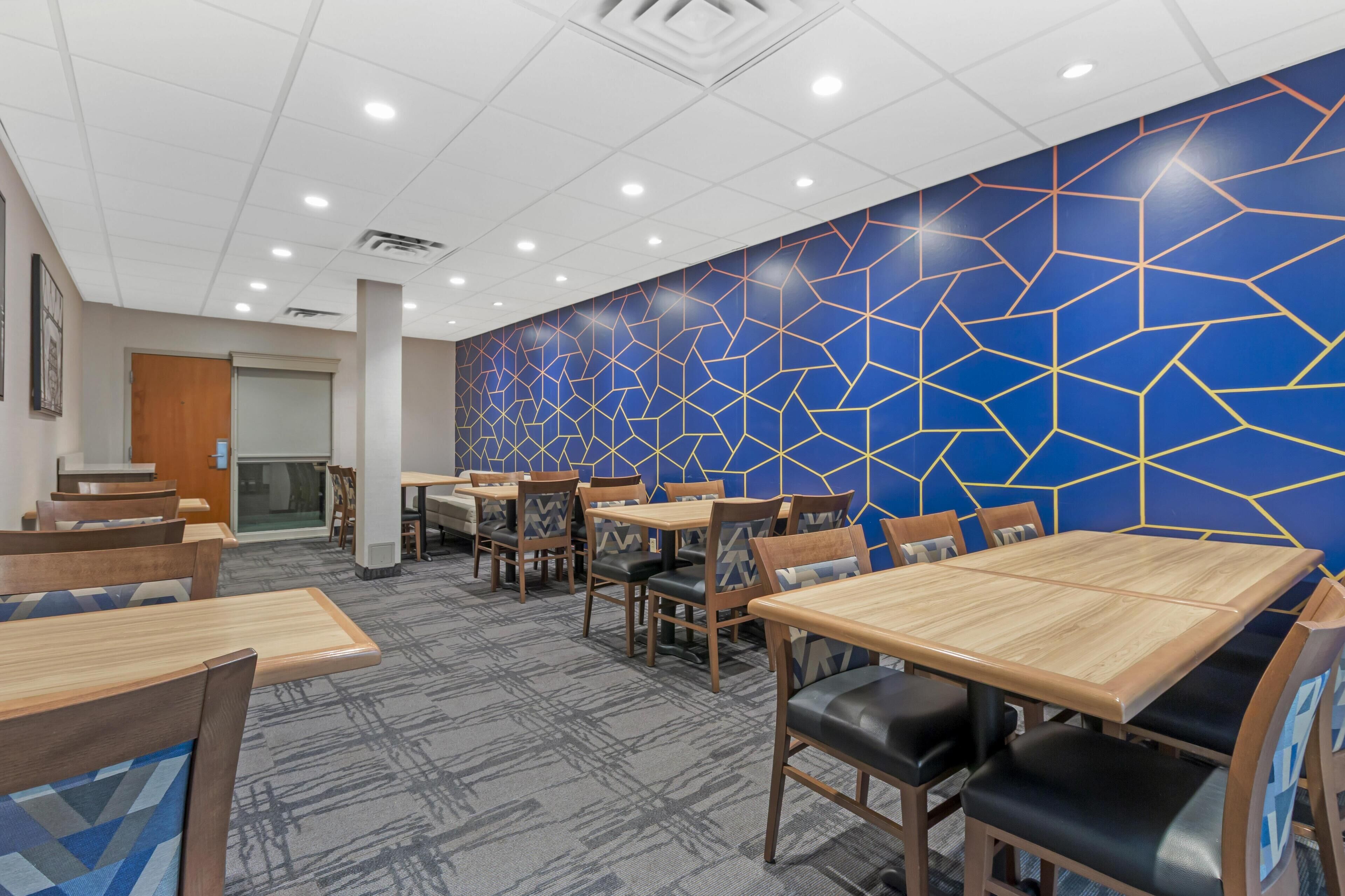 A hotel dining area with multiple tables and chairs, a patterned wall, and a carpeted floor.