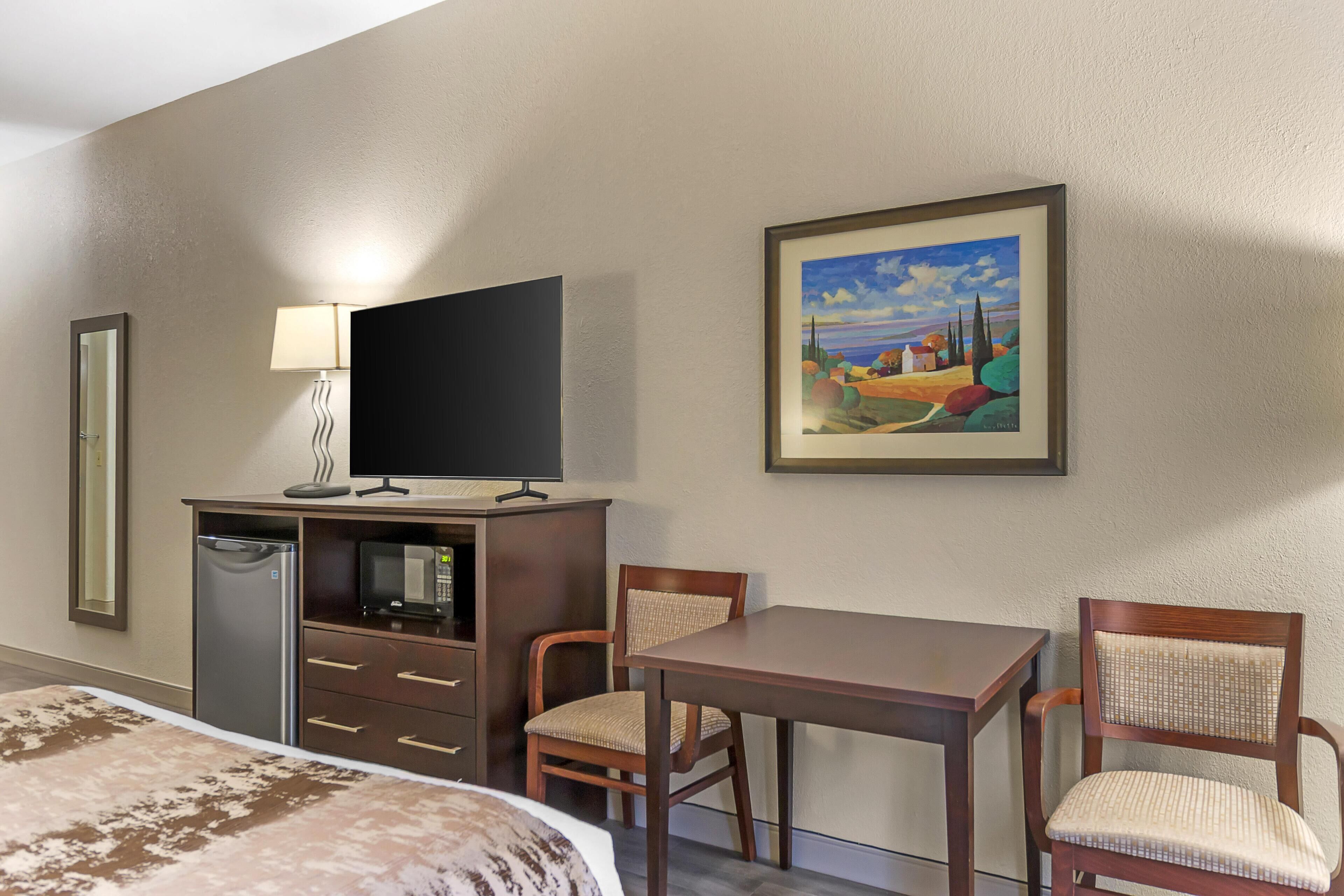 A hotel room with a bed, a desk with a computer, a chair, a small refrigerator, and a painting on the wall.