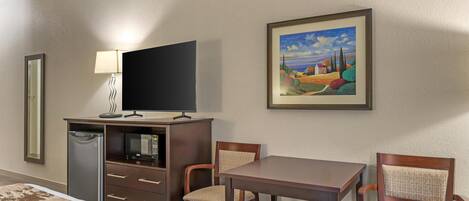 Habitación de hotel con cama, escritorio con computadora, silla, refrigerador pequeño y un cuadro en la pared.