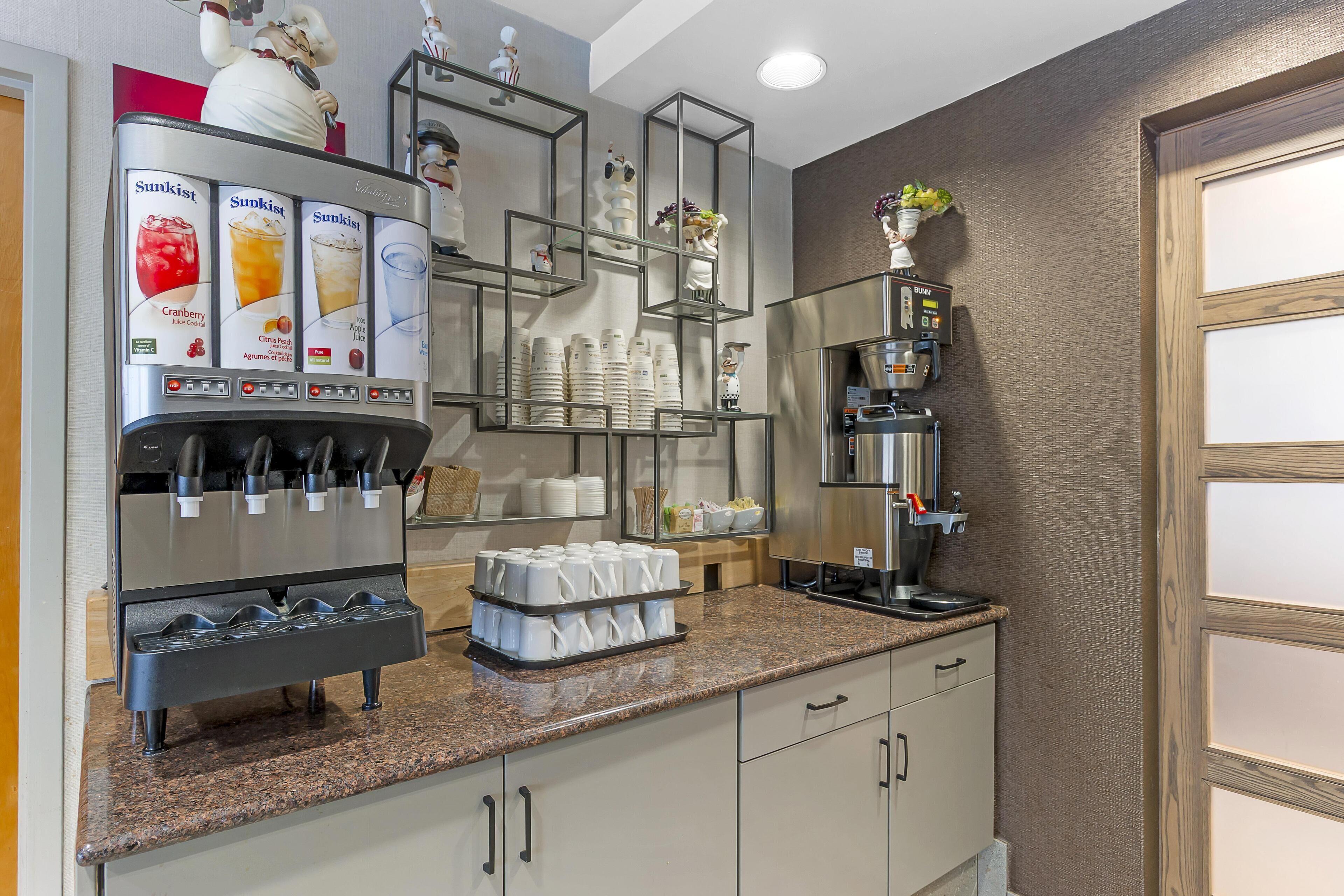 Una estación de autoservicio para bebidas con varias opciones, una máquina de café y tazas y platos blancos ordenados en la encimera.
