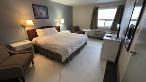 A hotel room with a bed, a desk, a chair, a small table, and a window. - Burntwood Hotel (Thompson)