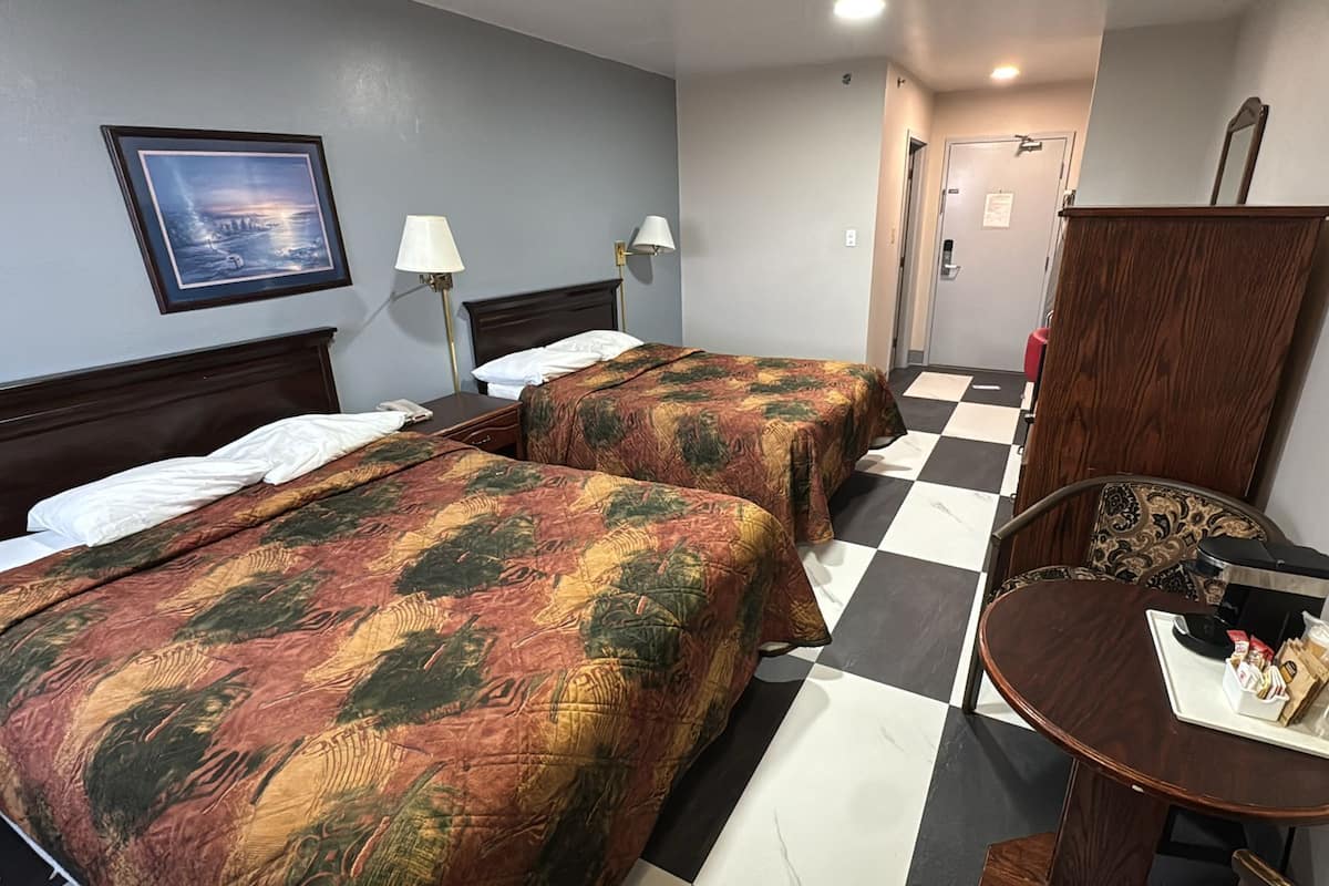 A hotel room with two beds, a checkered floor, a desk with a tray of items, and a framed picture on the wall.