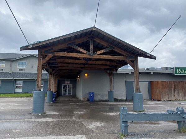 A covered outdoor area with a wooden roof structure, a door leading to a building, and a sign with the word "REG". - Burntwood Hotel (Thompson)
