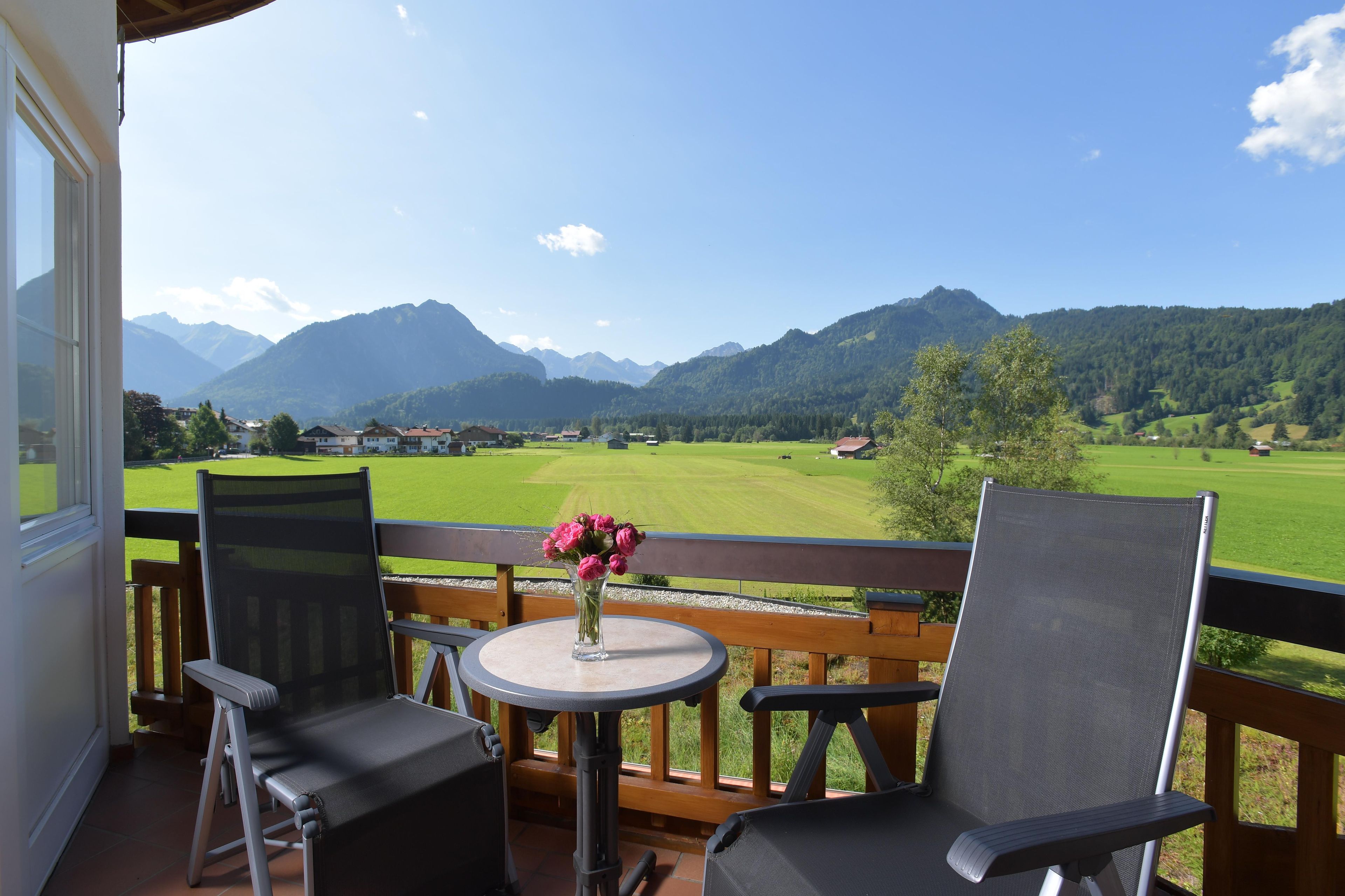 Un balcon avec deux chaises et une petite table, d’où l’on peut admirer un paysage verdoyant avec des montagnes à l’horizon.