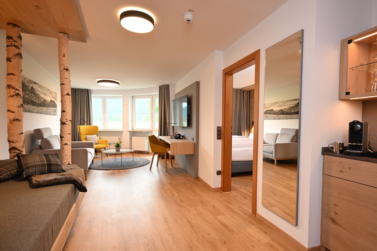 A modern hotel room with a wooden floor, a bed, a desk, a chair, a sofa, a TV, and a large mirror.