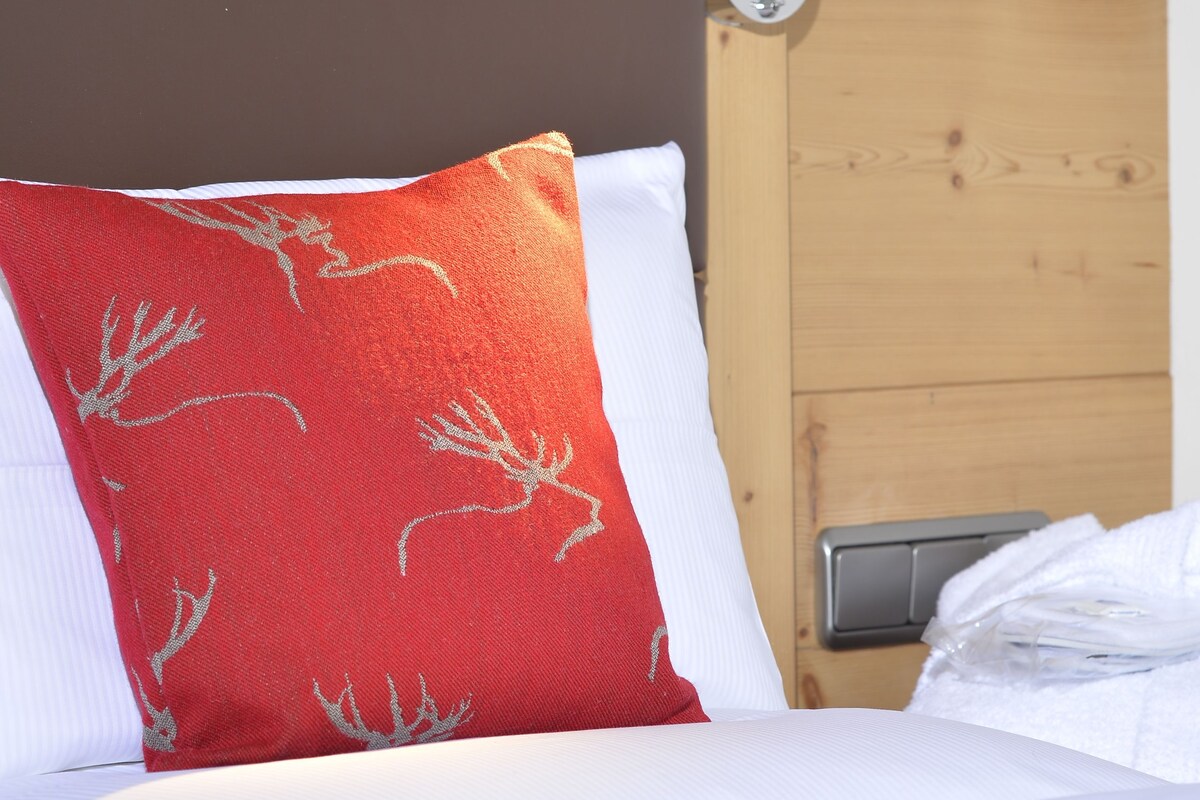A bed with white bedding, a red pillow with a tree pattern, and a headboard with a wooden panel and a wall-mounted lamp.