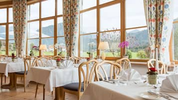 A restaurant with multiple tables set for dining, large windows with floral curtains, and a view of mountains.