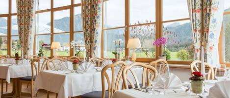Un restaurante con varias mesas preparadas para comer, amplias ventanas con cortinas florales y vistas a montañas.