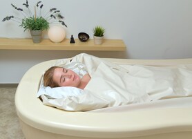 Una persona acostada en una cama con una manta blanca, un almohadón color beige y un cabecero color beige. Hay una estantería con una planta en maceta, una pequeña estatua y un cuenco decorativo.