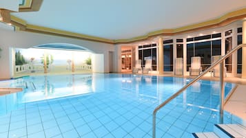 Ein Hallenbad mit hellblauen Fliesen, ein Blick ins Freie durch ein großes Fenster und eine Treppe, die zu einem Balkon führt.