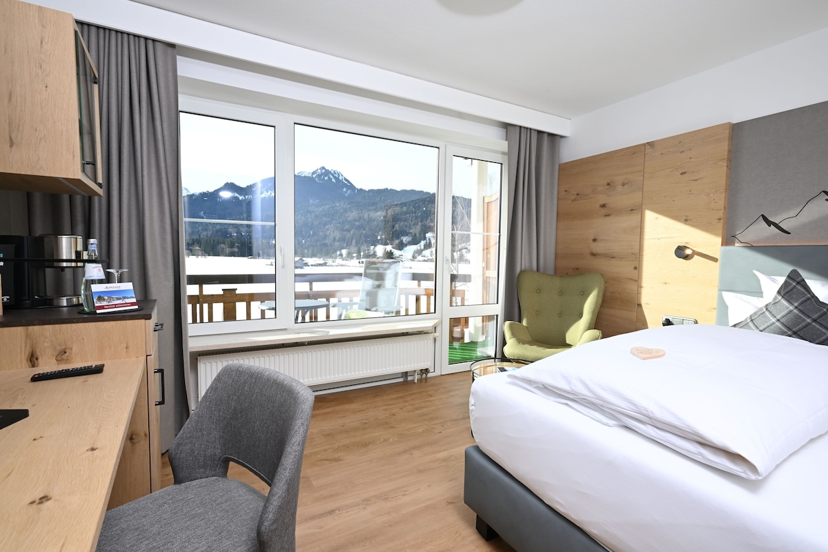 A hotel room with a bed, a desk, a chair, and a view of mountains through large windows.