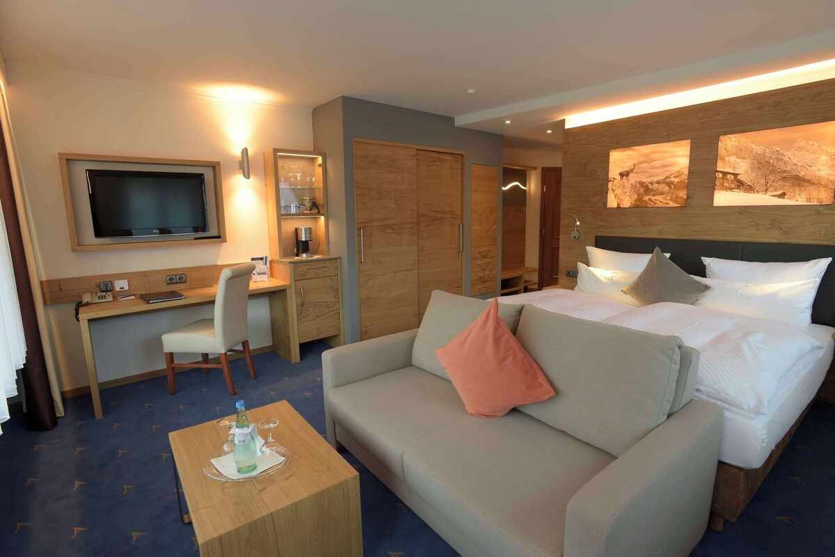 A hotel room with a bed, a sofa, a desk, a television, and artwork on the wall.