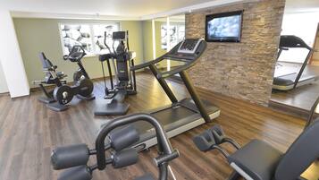 Academia com diversos equipamentos de exercício, incluindo esteiras e aparelhos de musculação, e uma televisão presa a uma parede de pedra.