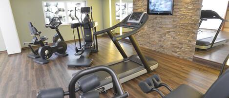 A gym with various exercise equipment, including treadmills and weight machines, and a television mounted on a stone wall.