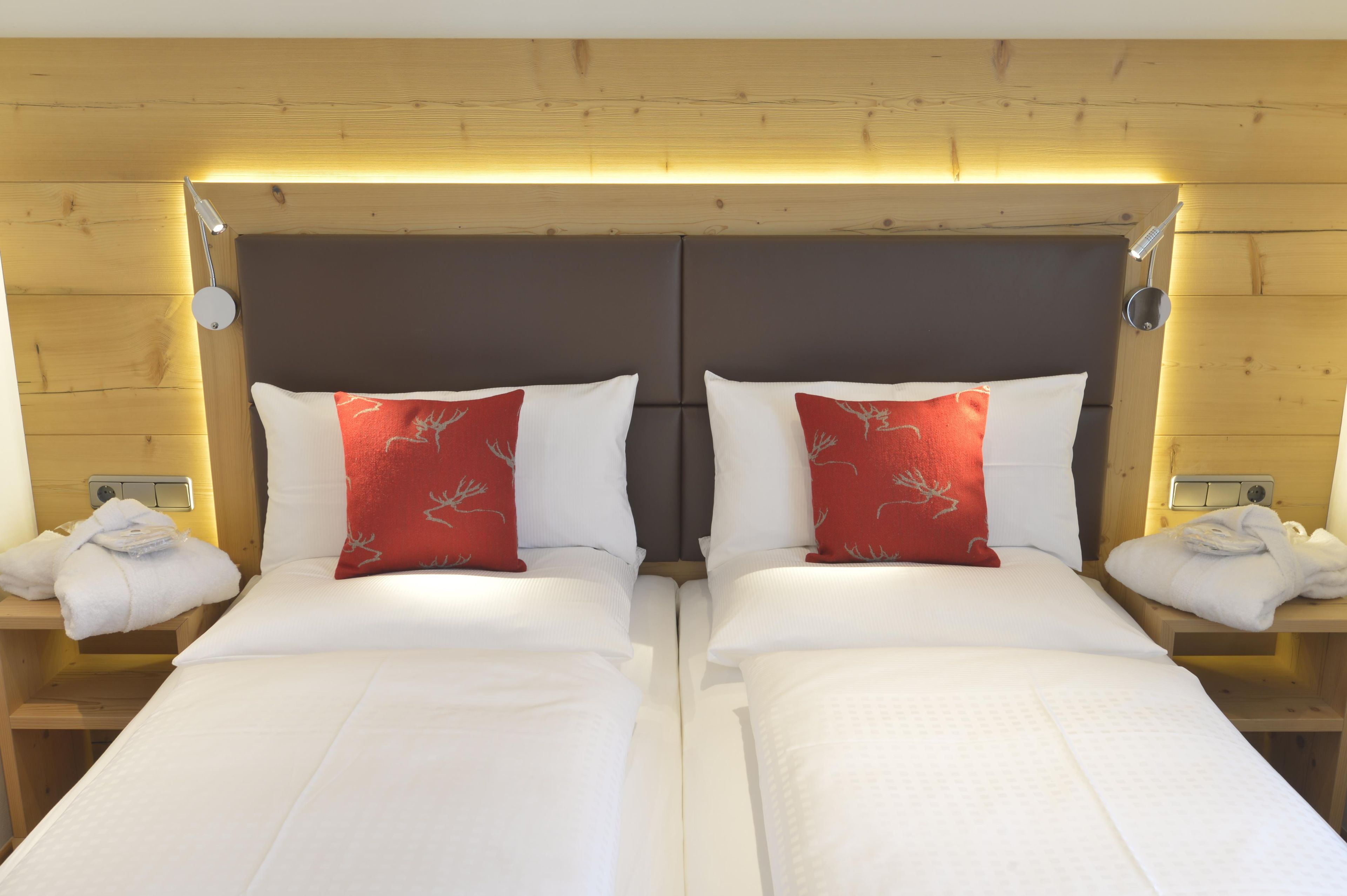A hotel room with two beds, each with a white mattress and a red pillow with a deer design. There are wooden headboards, nightstands with towels, and a wooden ceiling.