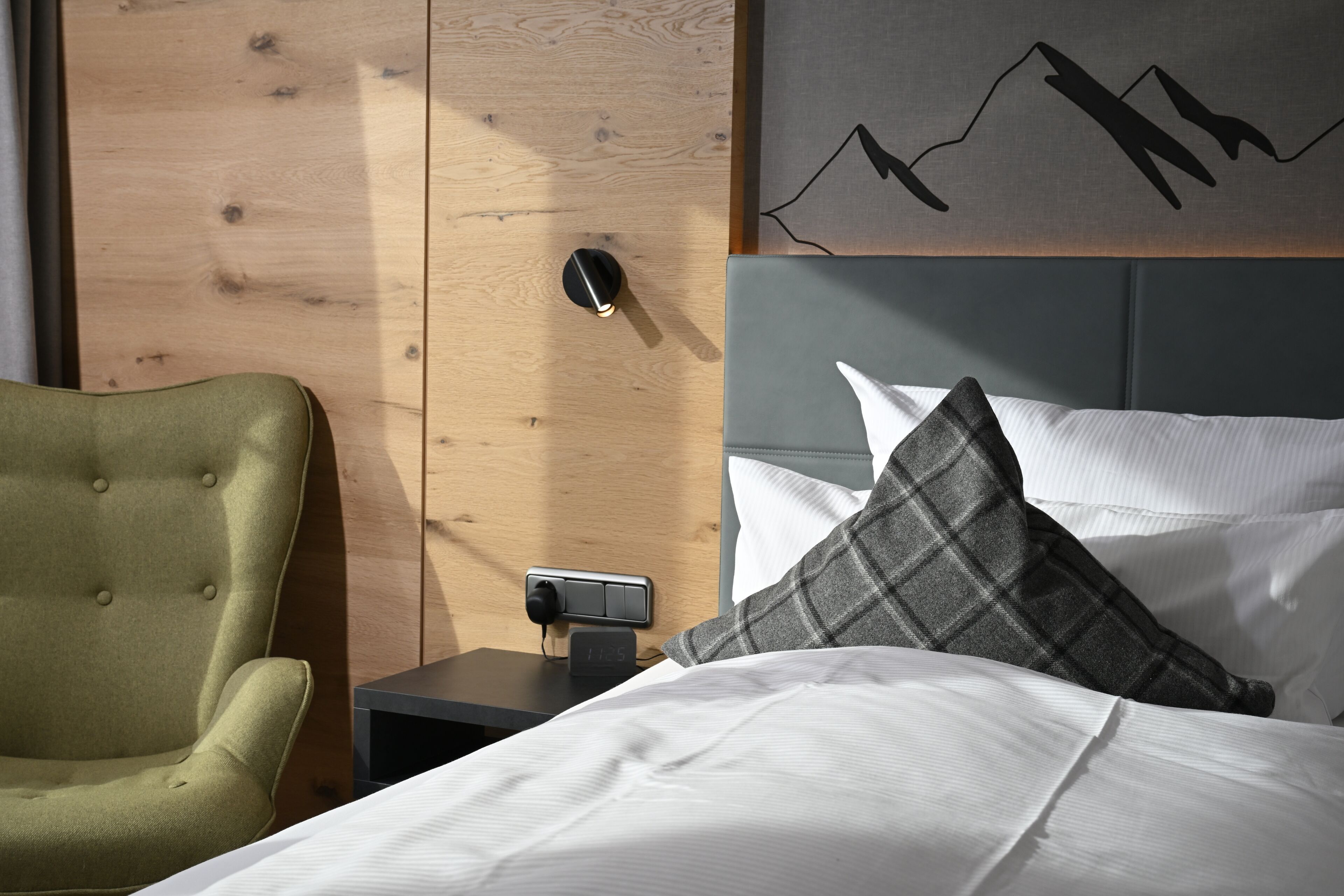 Une chambre d’hôtel avec un lit, une table de chevet, une horloge murale, une armoire en bois et un fauteuil vert.