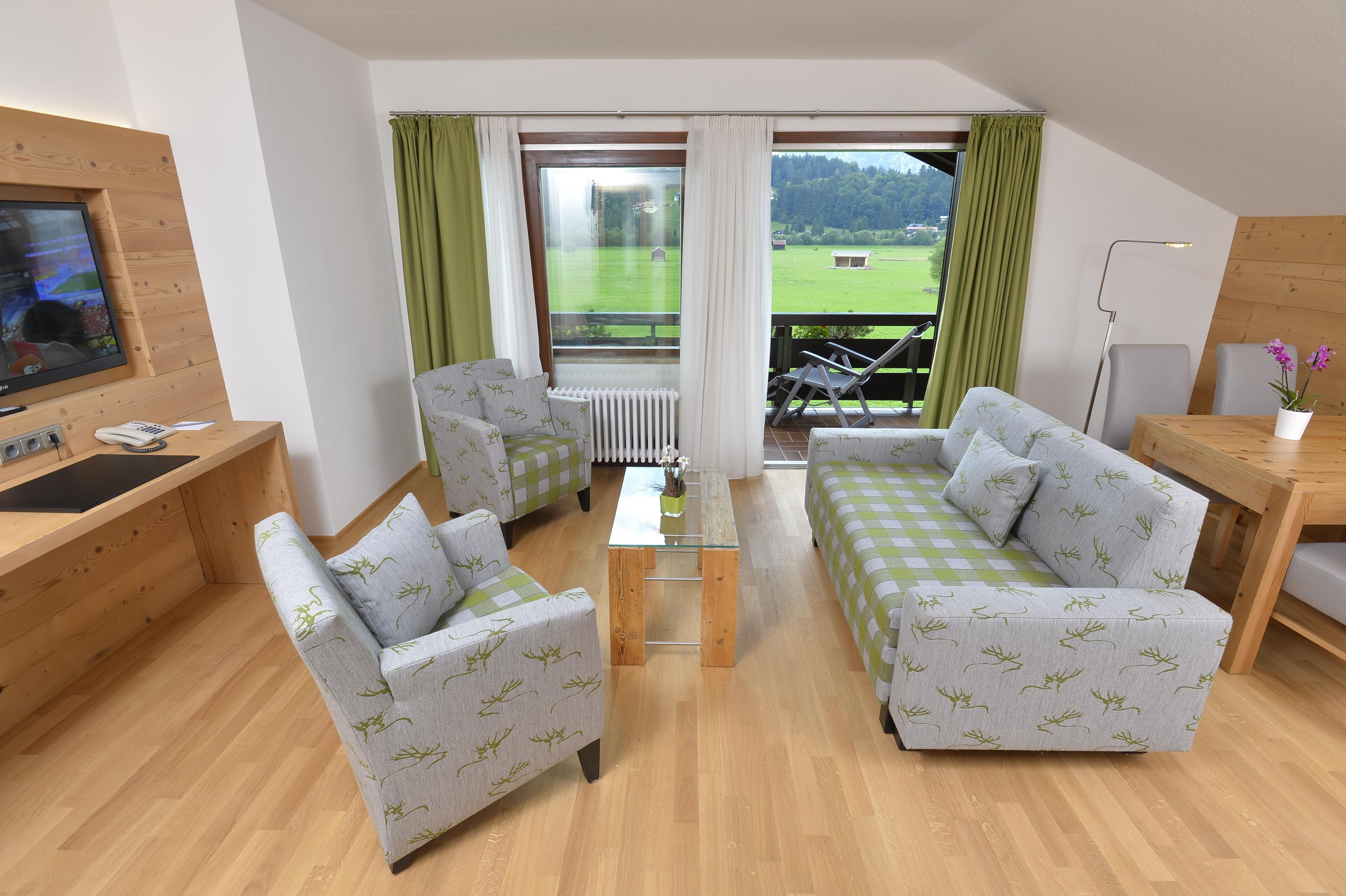 A modern living room with a sofa, chairs, a TV, and a balcony with a view of a green landscape.