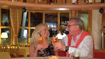 A couple toasting with drinks at a table in a restaurant with wooden furniture and large windows.