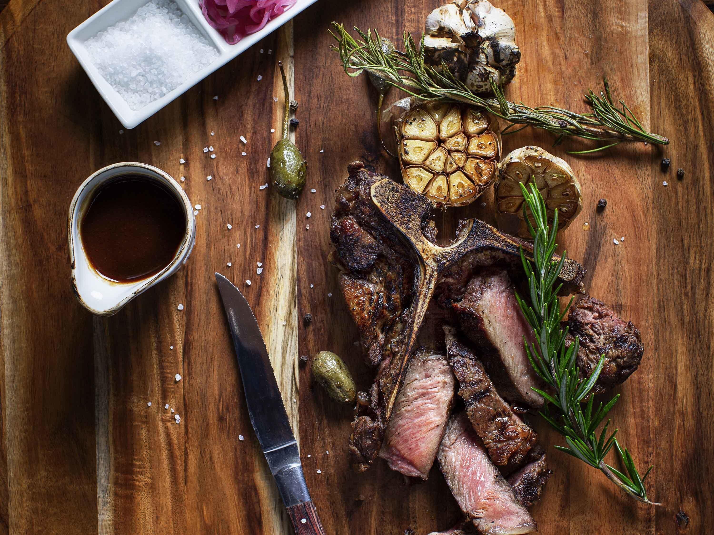 A steak with rosemary and garlic on a wooden table, a knife, and a small bowl of salt.