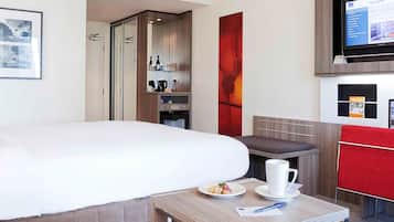 A modern hotel room with a large bed, a small table with a plate of food and a cup, a TV, and a red accent wall.