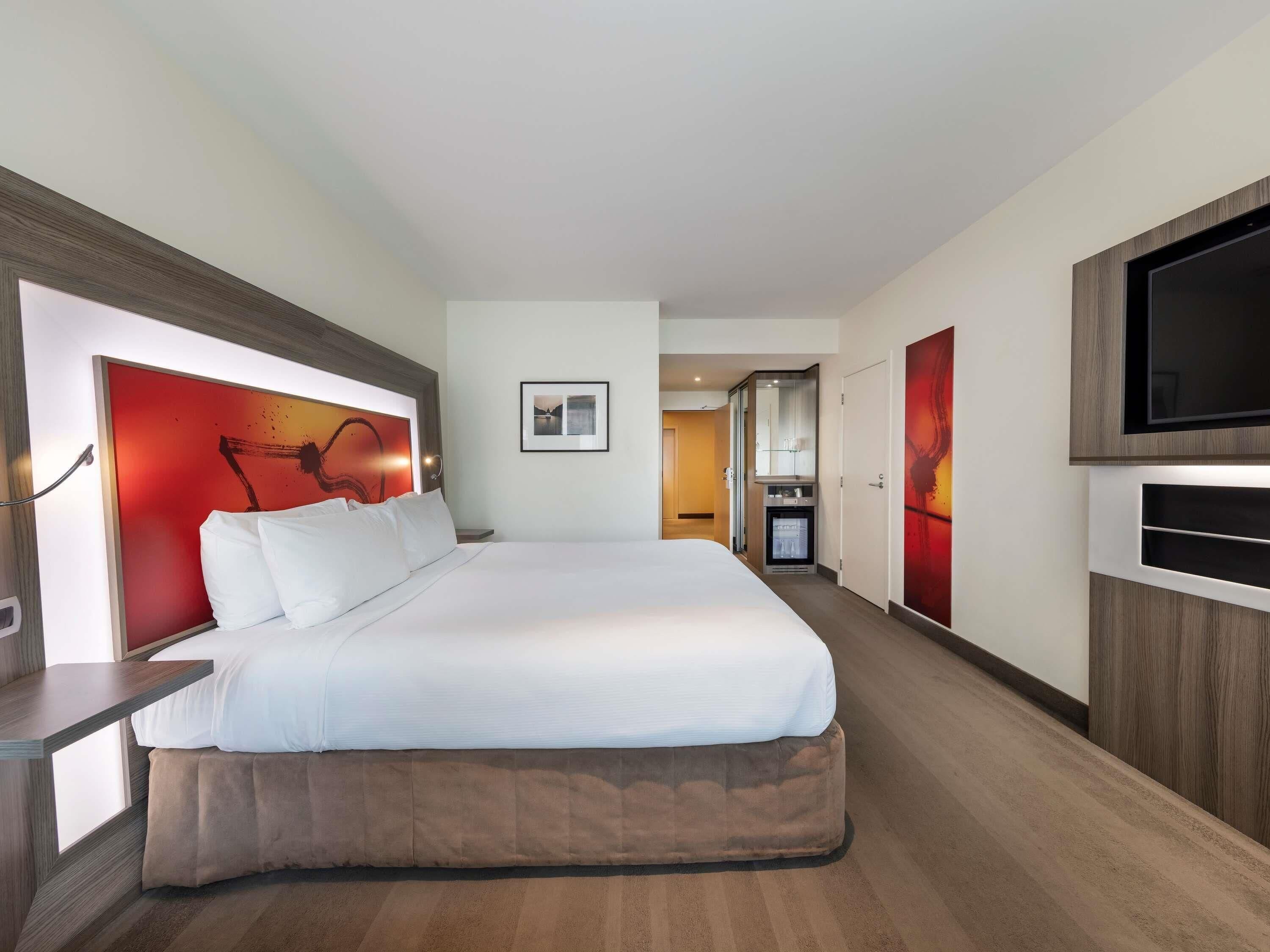 A hotel room with a large bed, a television mounted on the wall, and a minibar.