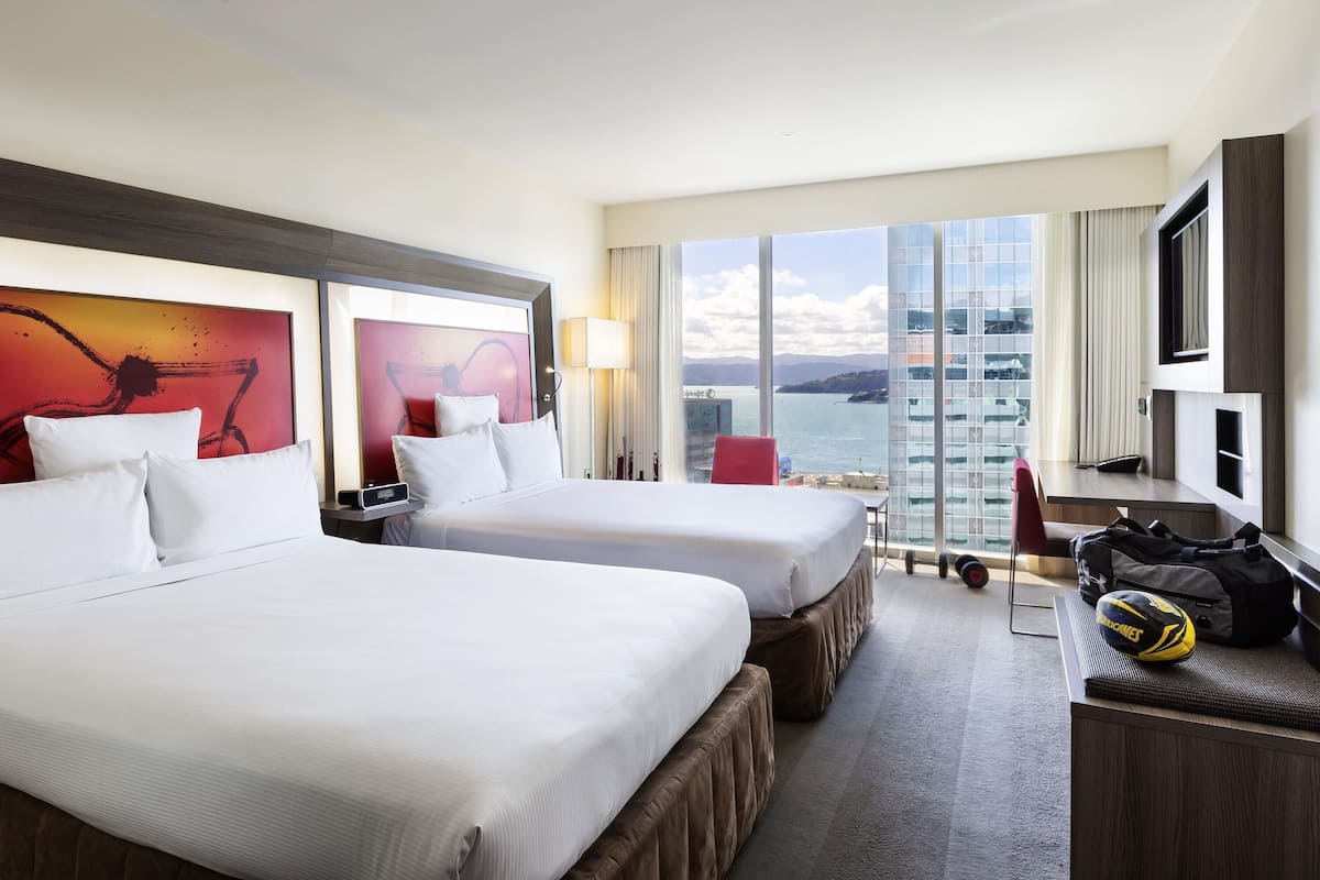 A hotel room with two beds, a desk, a chair, a TV, and a large window with a view of the sea.