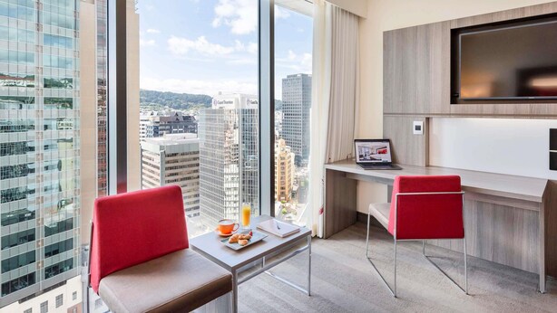 A modern hotel room with a large window offering a city view, a desk with a laptop, and two red chairs. - Novotel Wellington (Wellington)