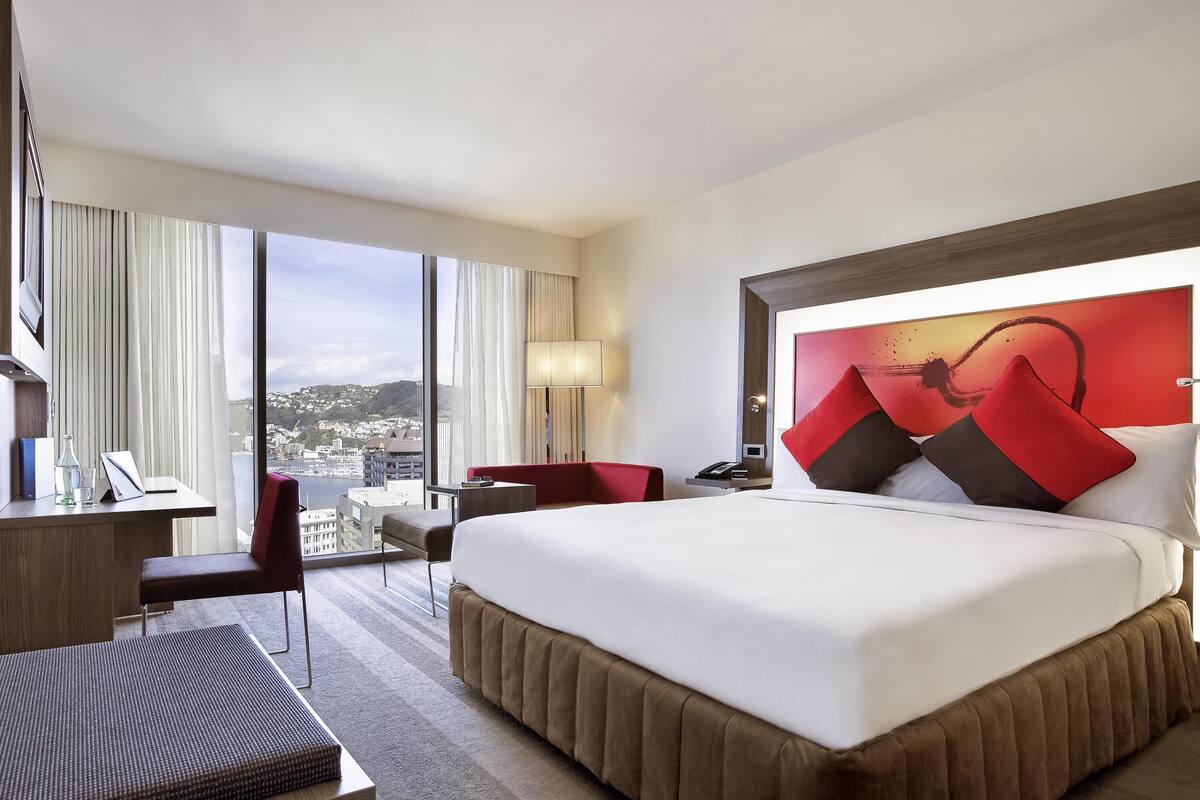 A hotel room with a large bed, a desk with a chair, a small table, and a view of a cityscape through large windows.