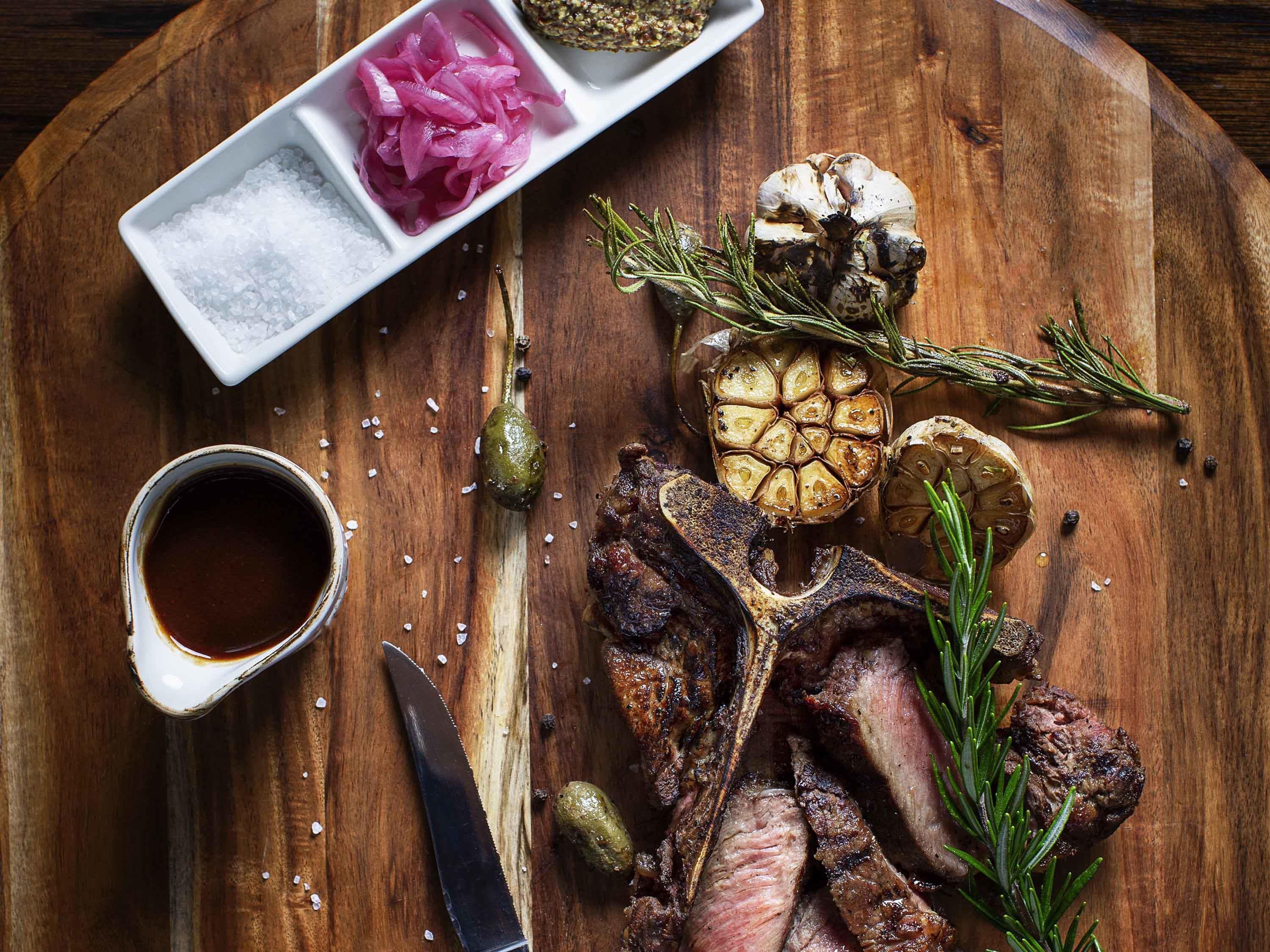 A wooden cutting board with a steak, garlic, rosemary, and a small dish of pickled onions.