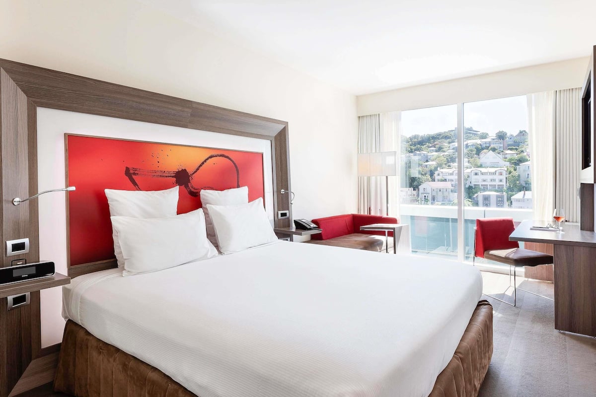A hotel room with a large bed, a red accent wall, a desk with a chair, and a view of buildings through large windows.