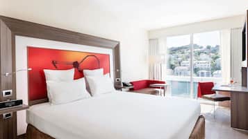 A hotel room with a large bed, a red accent wall, a desk with a chair, and a view of buildings through large windows.