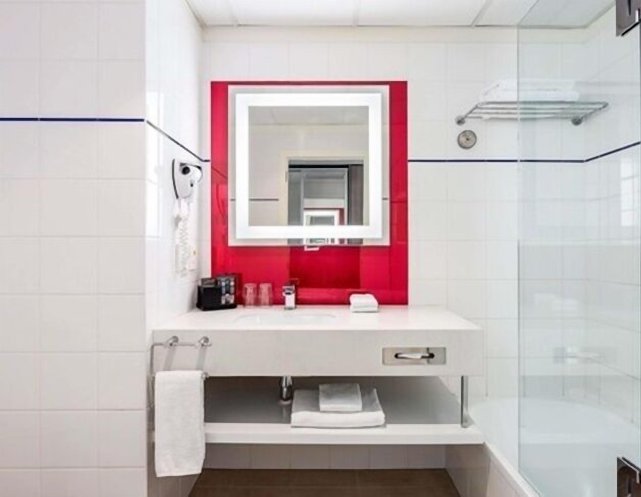 Une salle de bain moderne avec un meuble-lavabo blanc, un mur d’accent rouge, un grand miroir et une cabine de douche en verre.