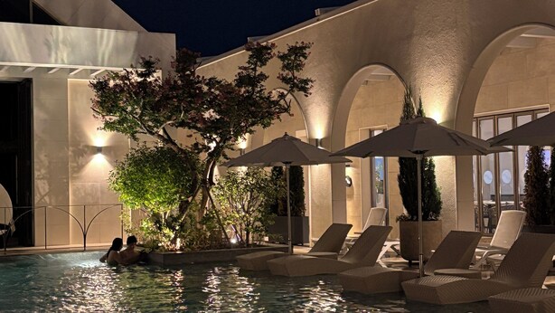 Una piscina al aire libre con sillas de descanso y sombrillas, rodeada por un edificio moderno con ventanas arqueadas y un árbol.