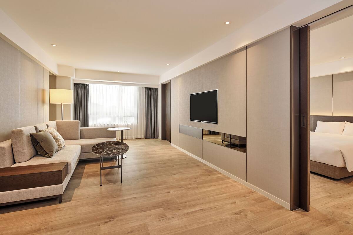 A modern hotel room with a sofa, a small round table, a TV mounted on the wall, and a bed.