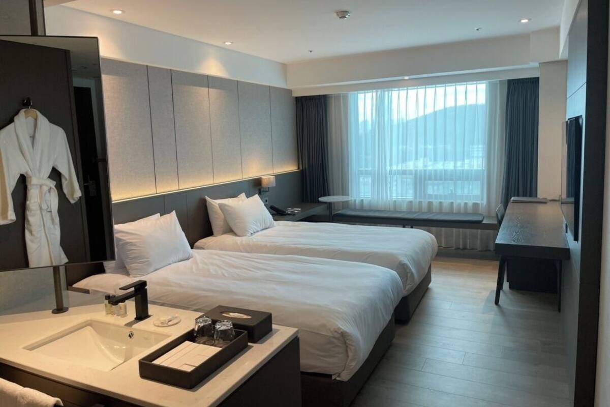 A modern hotel room with two beds, a large mirror, a vanity with a sink, and a view of the city through large windows.
