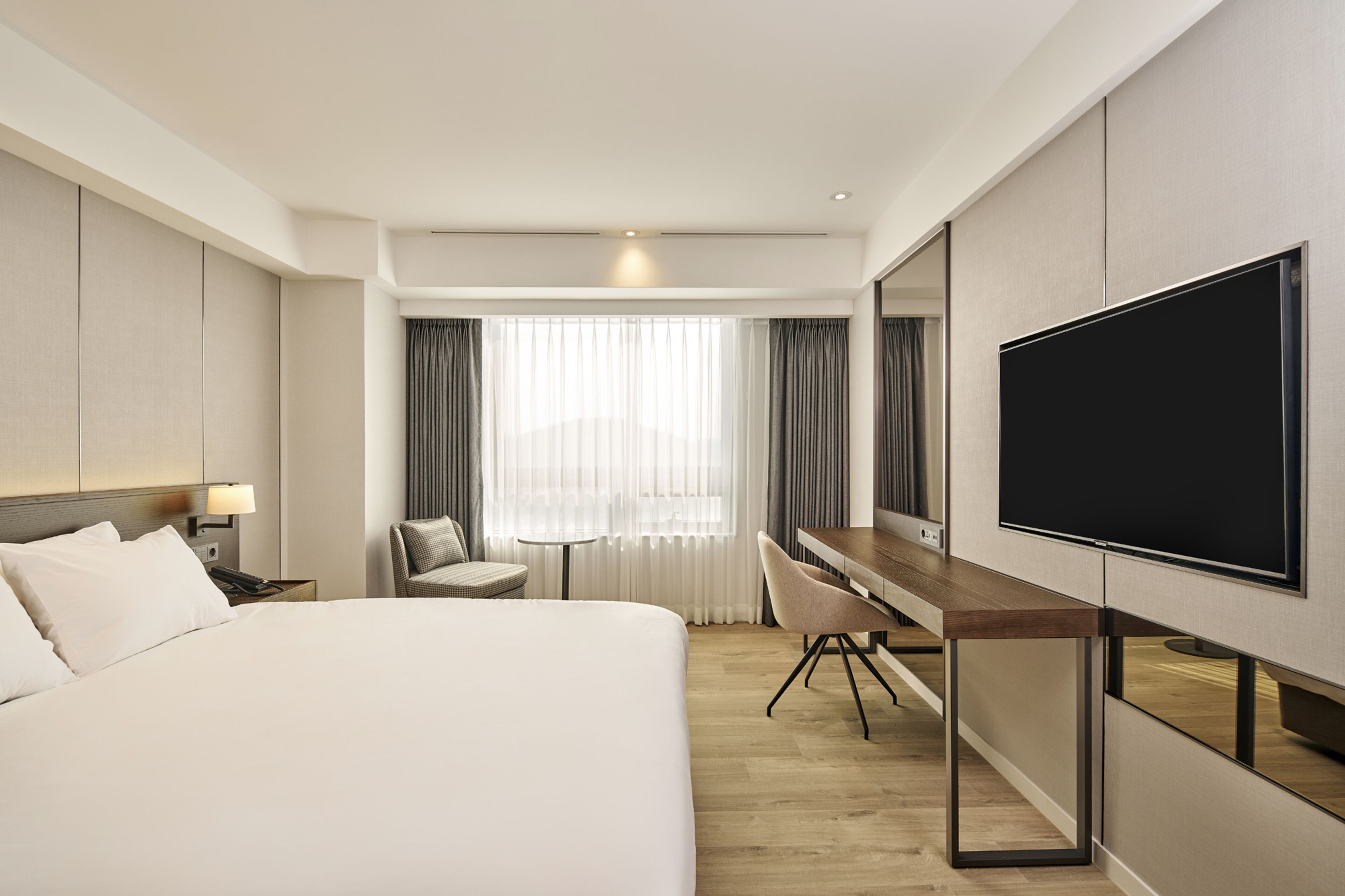 Ein Hotelzimmer mit einem großen Bett, einem Schreibtisch mit Stuhl, einem Flachbildfernseher und einem Fenster mit Vorhängen.