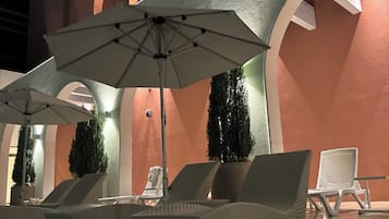 Zona de piscina con sillas de descanso y sombrillas, rodeada por un edificio con ventanas arqueadas y una pared con textura.